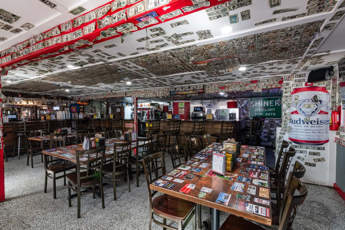 Eclectic bar and dining space decorated with dollar bills covering the ceiling and walls, featuring wooden tables with beer label designs, bar seating, and a laid-back dive bar atmosphere ideal for casual dining and drinks.