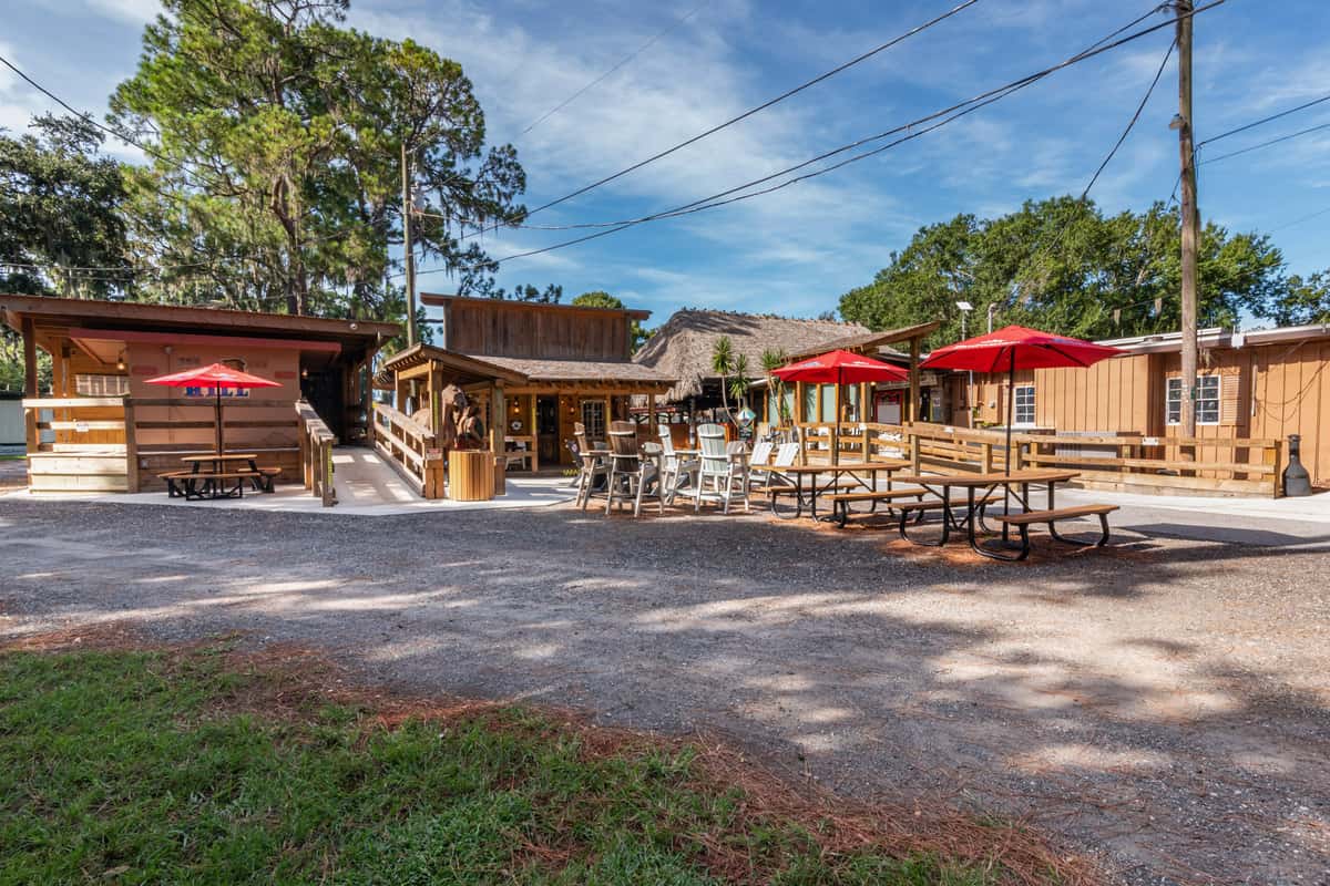 Outdoor view of a rustic wooden restaurant with picnic tables, red umbrellas, and a casual open-air seating area, surrounded by tall trees and tropical landscaping, evoking a laid-back, beachy dining experience.