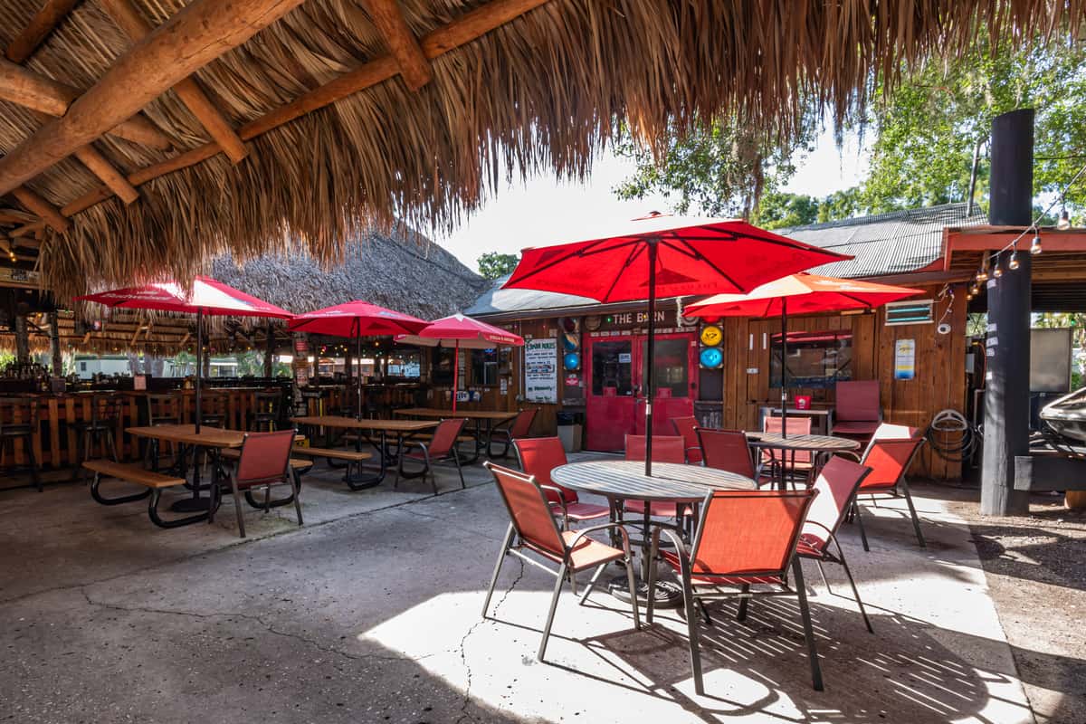 Covered tiki-style patio with red umbrellas and a mix of picnic tables and round patio seating, set against a rustic wooden bar backdrop, creating a relaxed tropical dining atmosphere ideal for enjoying food and drinks outdoors.