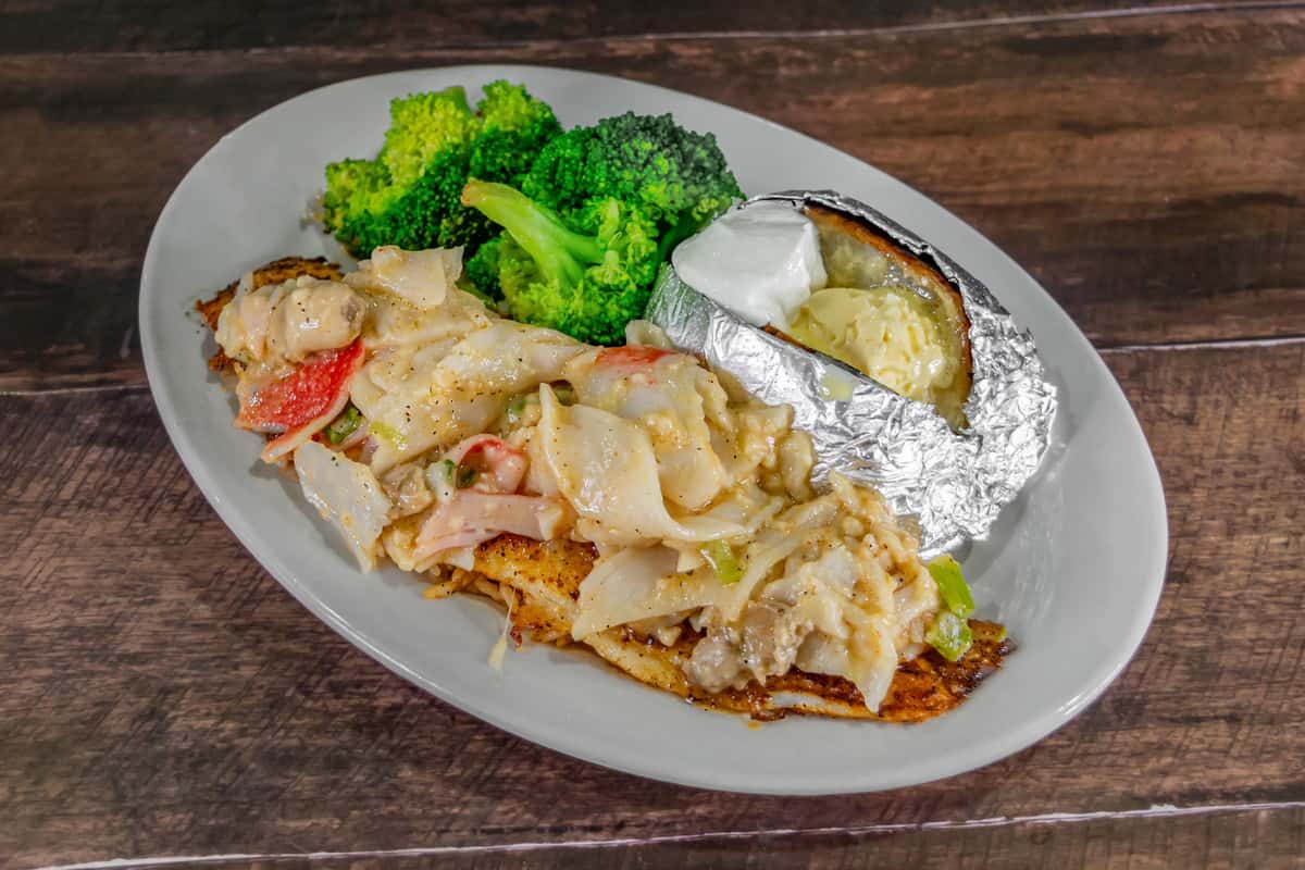 Blackened fish fillet topped with creamy seafood and vegetable medley, served with steamed broccoli and a foil-wrapped baked potato topped with butter and sour cream, showcasing a hearty coastal comfort dish.