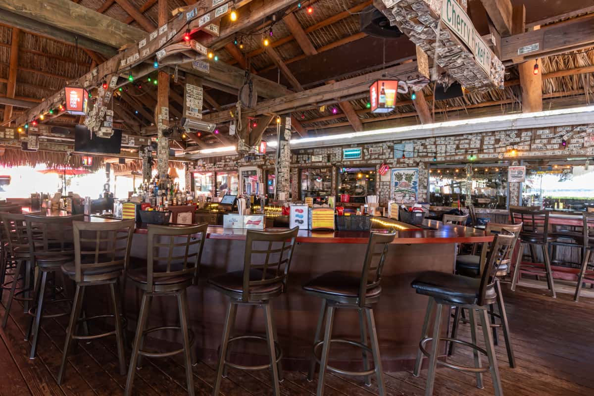 Island-themed bar with a thatched roof, wraparound counter seating, colorful string lights, and walls and beams covered in signed dollar bills, capturing a lively tropical vibe perfect for casual drinks and coastal dining.