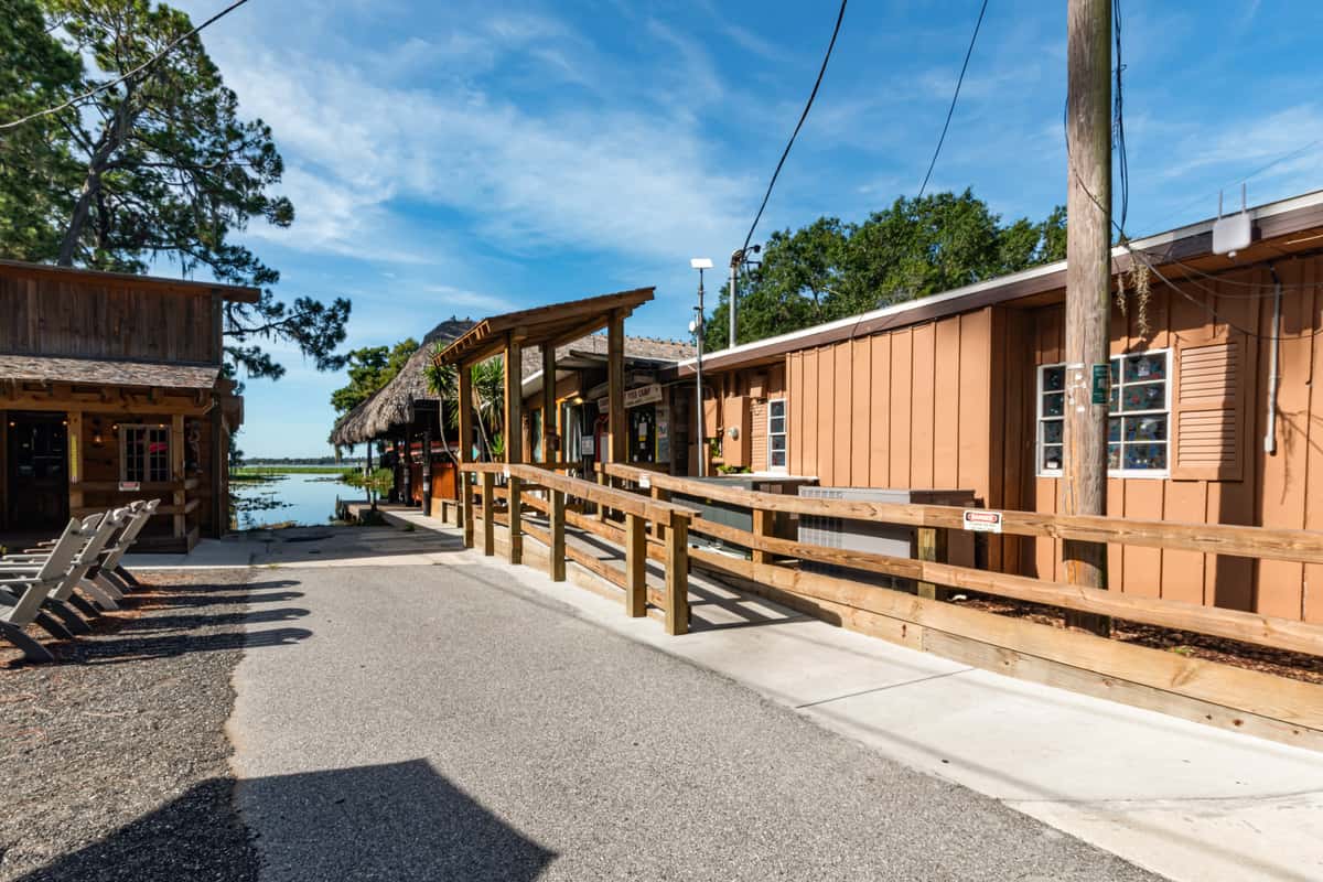 Sunny walkway between rustic wooden buildings at a lakeside restaurant, featuring a ramp with railings and a thatched-roof dining area in the distance, creating a welcoming and accessible waterfront dining atmosphere.
