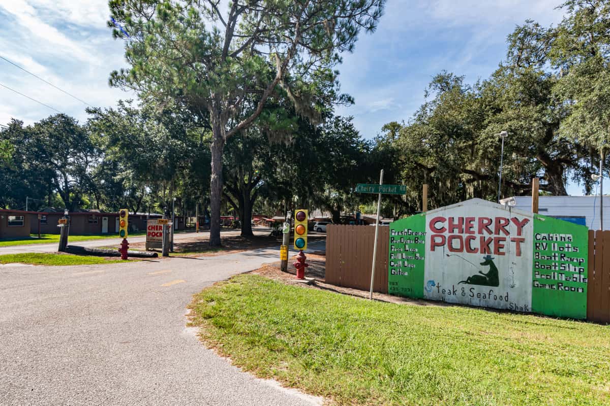 Entrance to Cherry Pocket Steak & Seafood Shak, marked by a rustic roadside sign listing amenities like cabins, RV lots, boat ramp, and game room, surrounded by large oak trees and set in a laid-back, wooded lakeside setting.