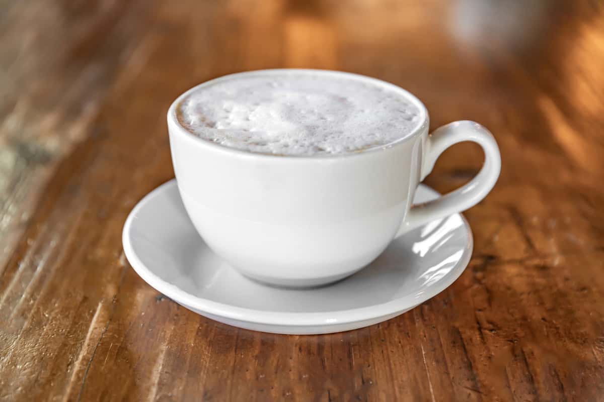 A frothy cappuccino on a white saucer in a coffee cup sits atop a rustic wooden counter.