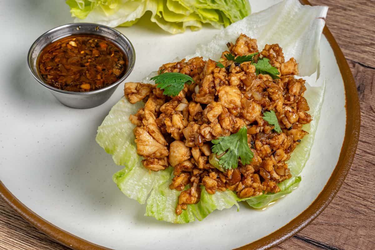 Minced chicken lettuce wrap topped with cilantro, served with a side of soy-based dipping sauce on a white plate.