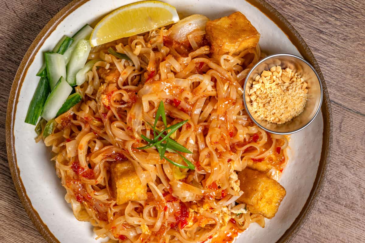 Tofu pad thai topped with scallions, served with crushed peanuts, cucumber sticks, and a lemon wedge on the side.