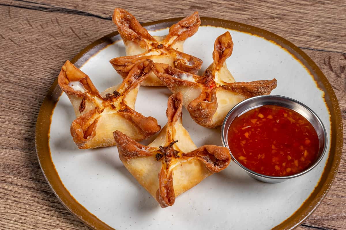 Golden crab rangoon served on a plate with a side of sweet chili dipping sauce.