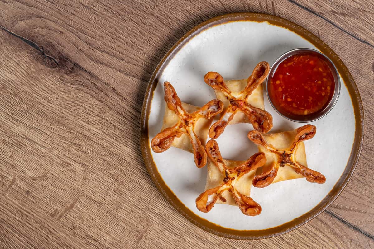 Golden crab rangoon served on a plate with a side of sweet chili dipping sauce.