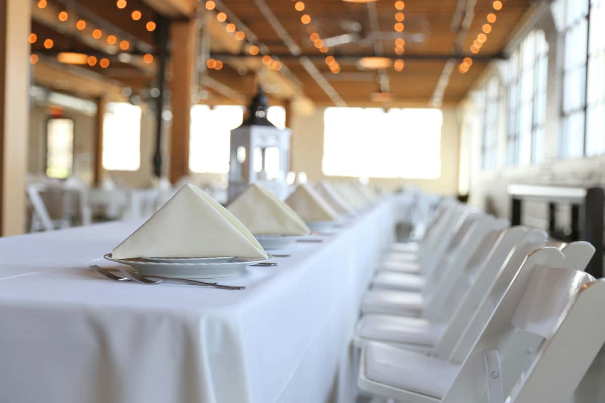Elegant event setting featuring a long table with white linens, folded napkins, and white chairs in a sunlit industrial loft with string lights.
