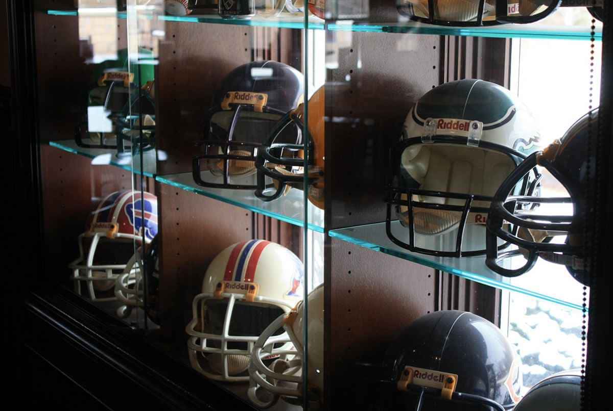 Football helmets displayed on glass shelves in a wooden cabinet, featuring various team designs and colors.