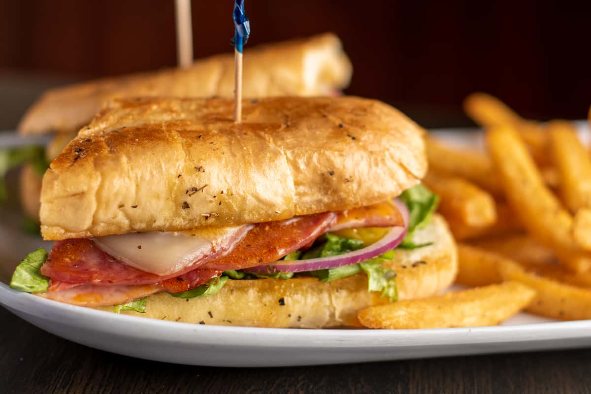 A toasted sandwich with layers of sliced deli meat, melted cheese, lettuce, tomato, and red onion, secured with a toothpick and served with a side of thick-cut fries on a white plate.