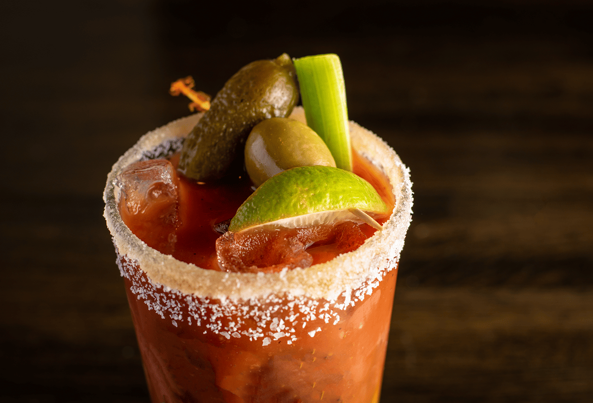 A Bloody Mary cocktail in a salt-rimmed glass, garnished with a lime wedge, green olives, a pickle, and a celery stick, served over ice on a wooden table.