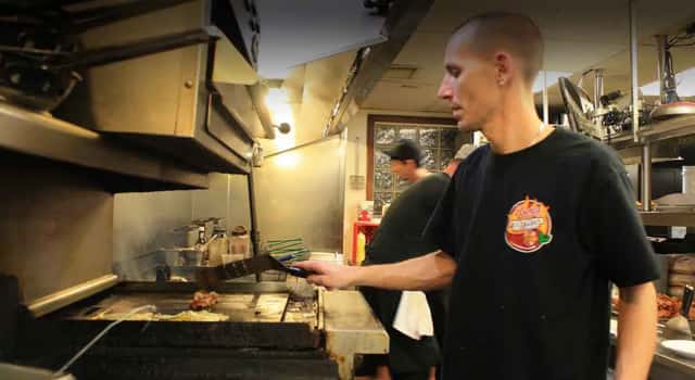 Cook grilling food on a flat-top in a restaurant kitchen, flipping meat with a spatula under stainless steel vents.