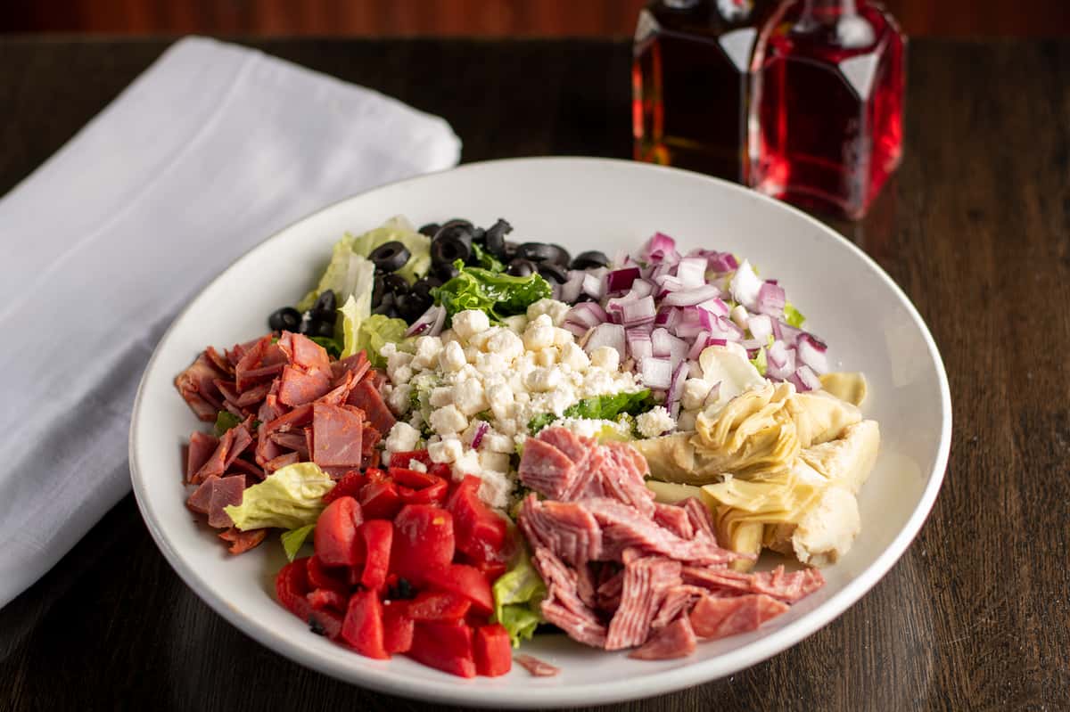 Antipasto salad with salami, ham, red peppers, olives, red onion, artichokes, and feta over lettuce in a white bowl.
