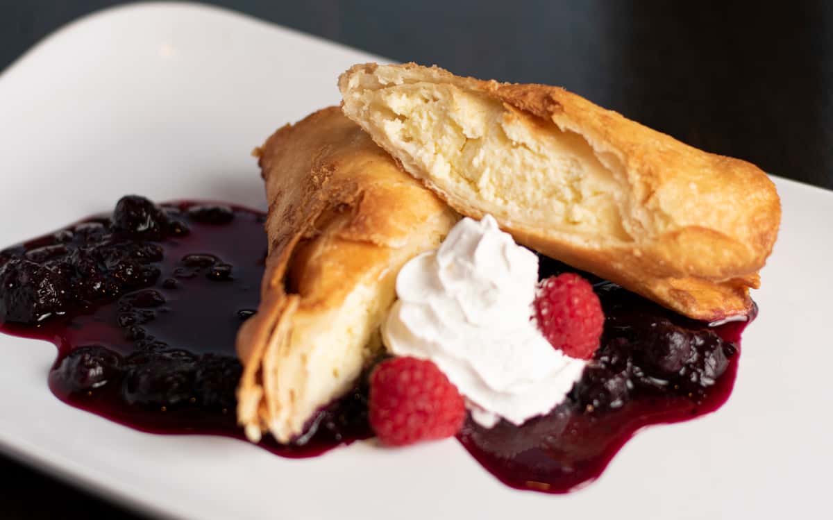 Slice of cheesecake-filled pastry topped with berry compote, whipped cream, and fresh raspberries on a white plate.
