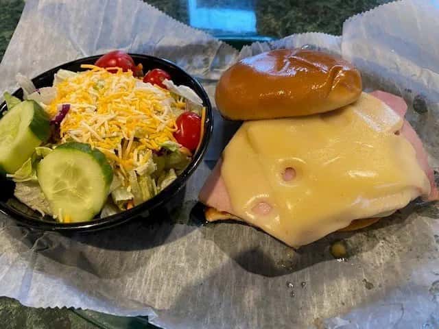 Classic ham and Swiss sandwich on a soft bun served with a side salad of mixed greens, cherry tomatoes, shredded cheddar cheese, and sliced cucumbers in a black bowl — perfect for a light American diner lunch.