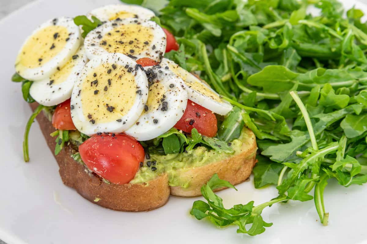 Avocado toast topped with sliced boiled eggs, cherry tomatoes, and black pepper, served with a side of fresh arugula.