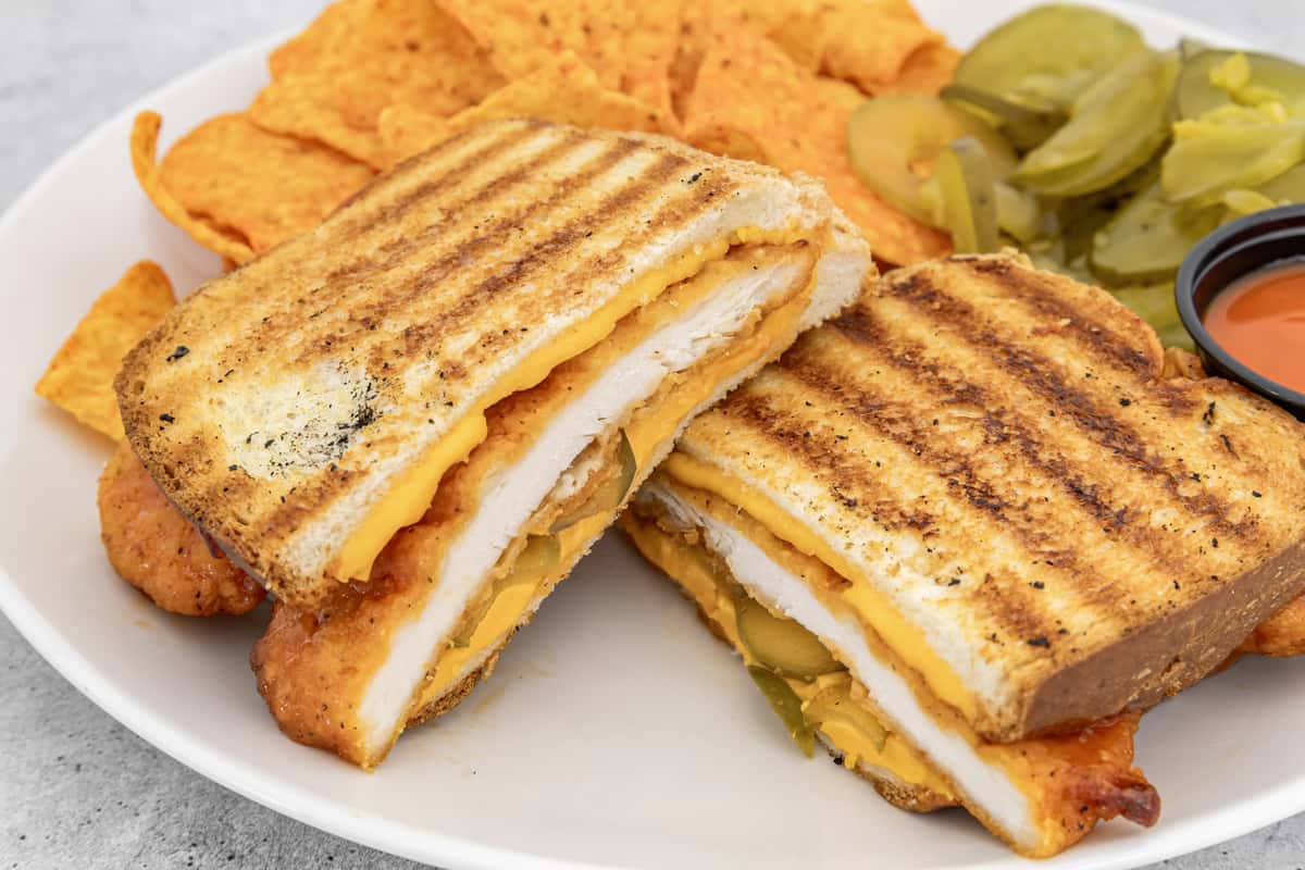 Crispy chicken grilled cheese with pickles and cheddar on toasted bread, served with nacho chips and a side of hot sauce.