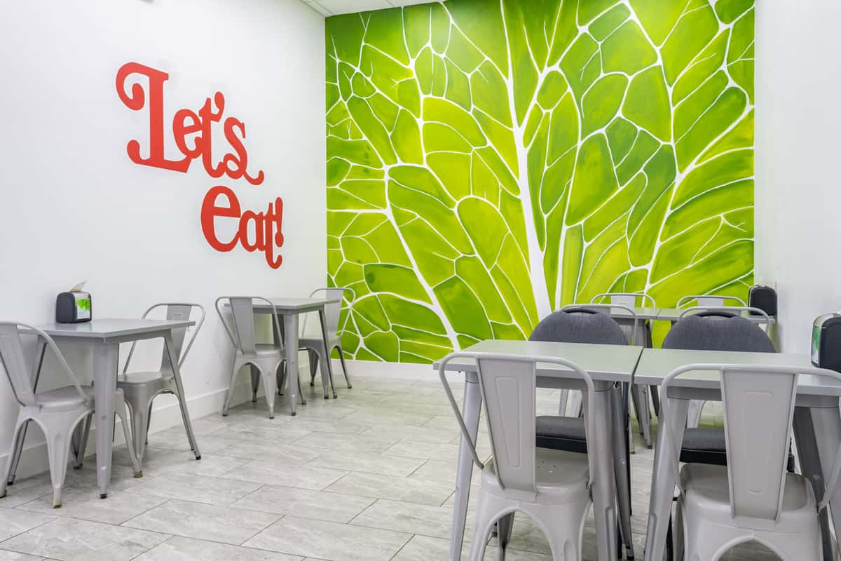 Modern dining area with gray metal chairs, green leafy wall art, and bold red "Let's Eat!" lettering on a white wall.