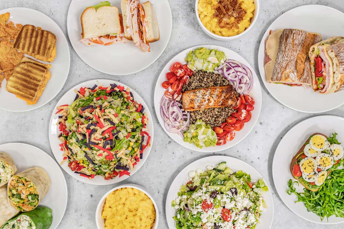 Assorted lunch dishes including paninis, deli sandwiches, mac and cheese, salads, wraps, and a quinoa bowl with grilled salmon.