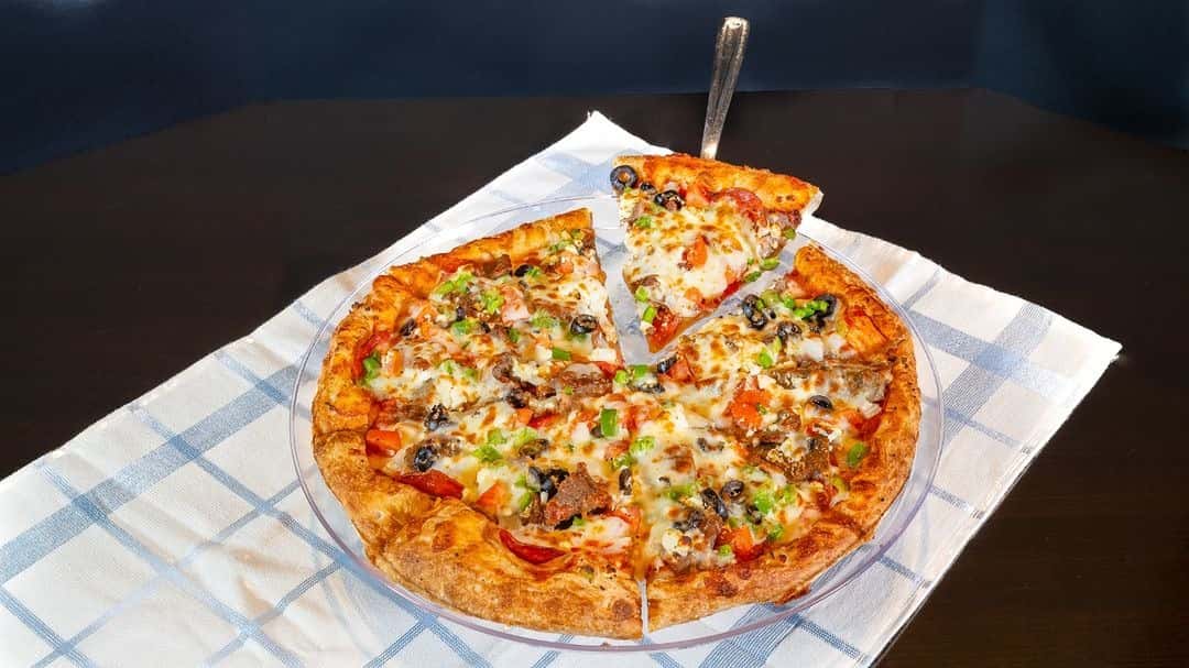 Supreme pizza on a clear plate with one slice lifted, topped with olives, peppers, tomatoes, sausage, and melted cheese.