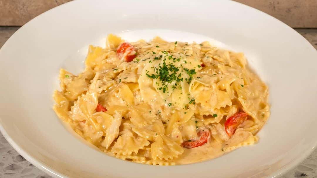 This is a dish of creamy bowtie (farfalle) pasta with visible cherry tomatoes and what appears to be shredded chicken, topped with grated cheese and chopped parsley.