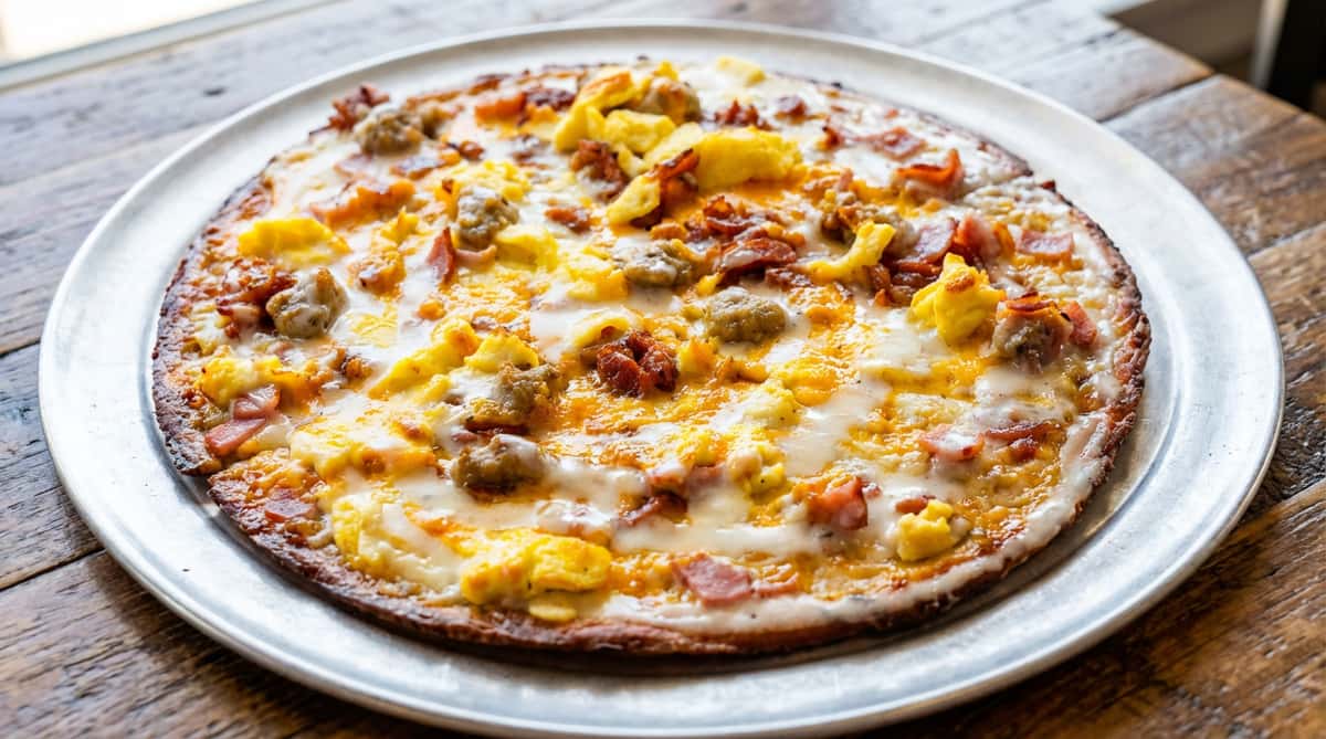 Breakfast pizza with scrambled eggs, sausage, bacon, ham, and melted cheese on a metal pan.
