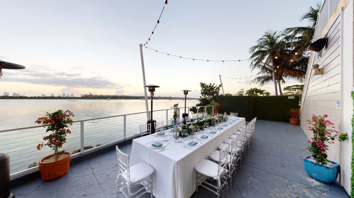 Outdoor waterfront event dining setup with long reception table, string lights, and sunset bay views