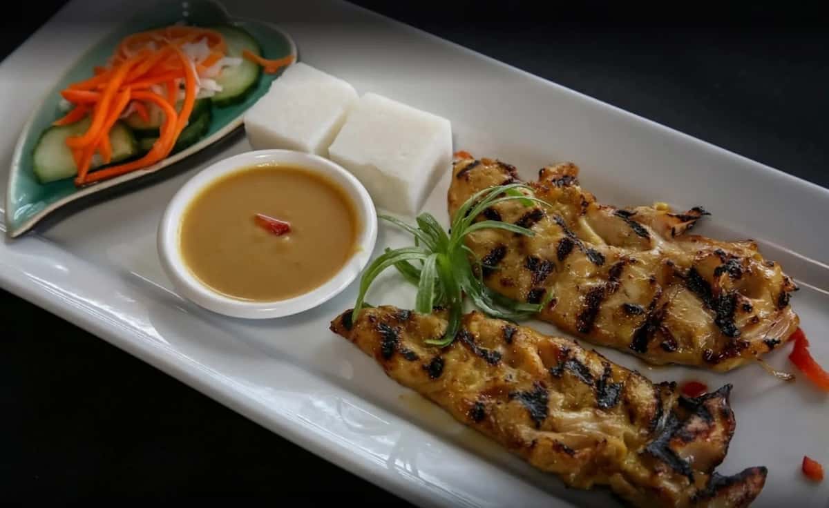 Grilled meat skewers served with a small bowl of peanut dipping sauce and white rice cakes