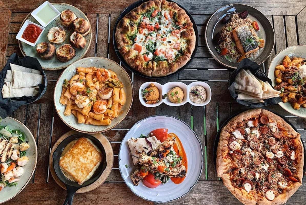 Rustic wooden table filled with a Mediterranean and Italian feast, featuring shrimp rigatoni pasta, wood-fired spinach and tomato pizza, pepperoni and sausage pizza, grilled chicken skewers with rice and pita, spanakopita in cast iron, seared salmon with farro and beets, saucy gnocchi with braised meat, baked garlic knots with dipping sauces, hummus trio, and fresh salad with shrimp and croutons.