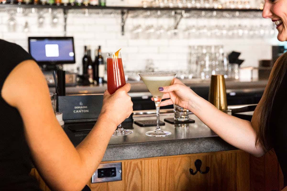 Two women clinking vibrant cocktails—one red and one pale green—at a stylish bar with a backdrop of hanging glassware and spirits.