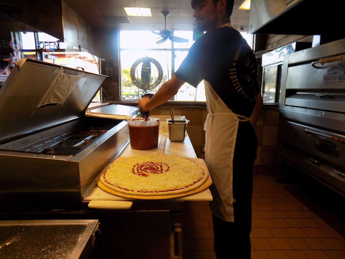 pizza chef spreading sauce on large cheese pizza in a busy pizzeria kitchen
