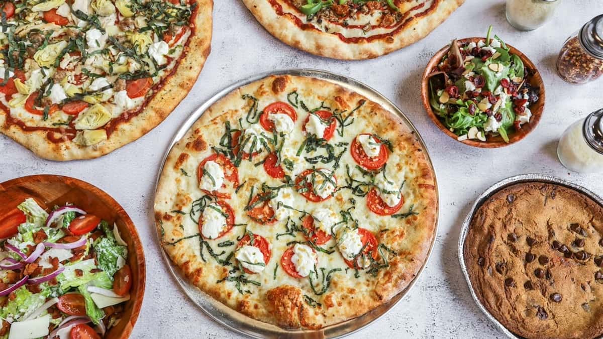 Overhead view of assorted pizzas with fresh salads and a chocolate chip cookie on a restaurant table