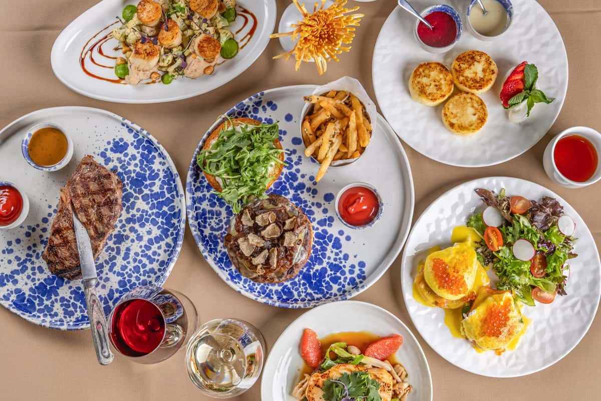 Flat lay of assorted gourmet dishes including steak, scallops, eggs Benedict, truffle burger, salads, and desserts with wine.