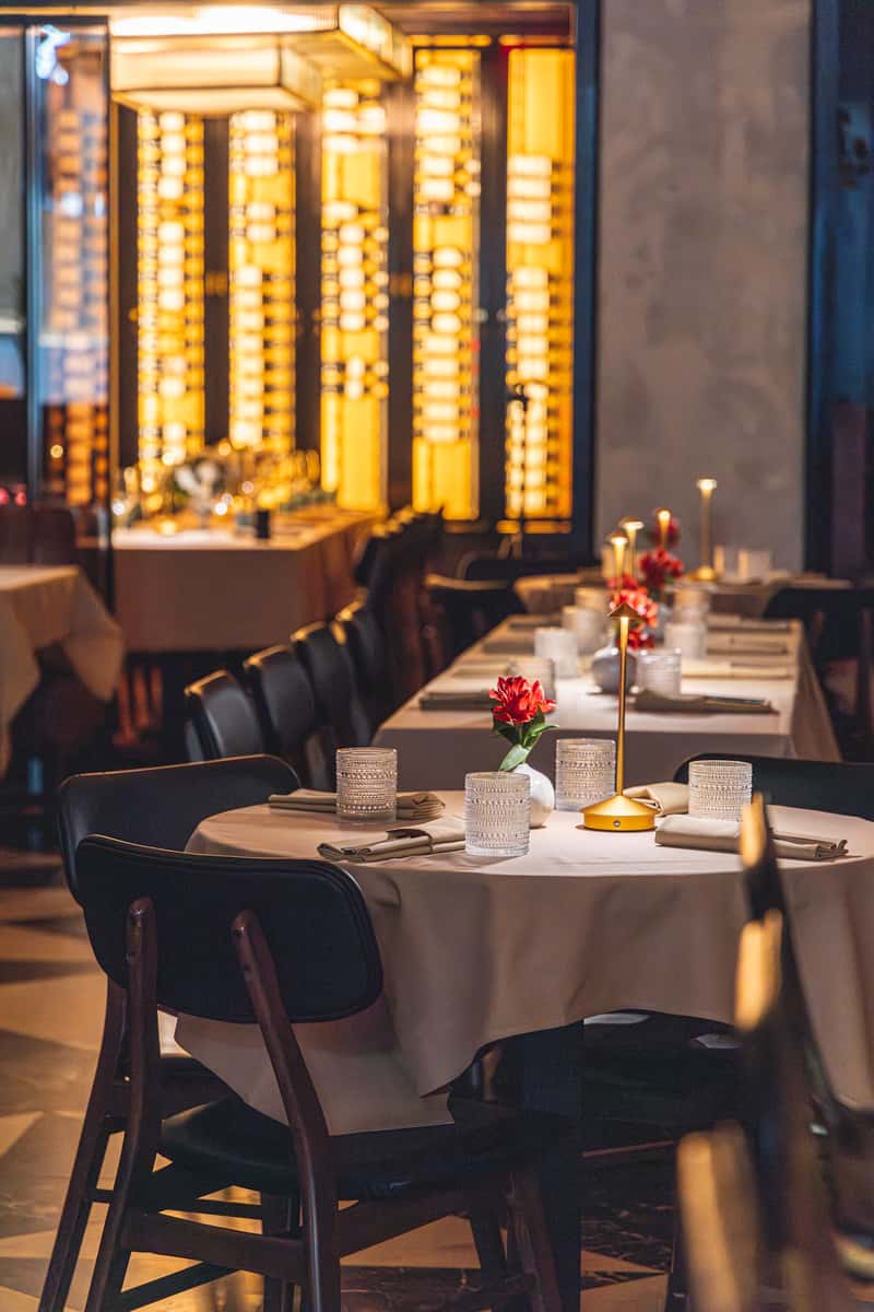 Private dining room with a long table set with wine glasses, napkins, yellow flowers, and blue candle holders beside a wine wall.