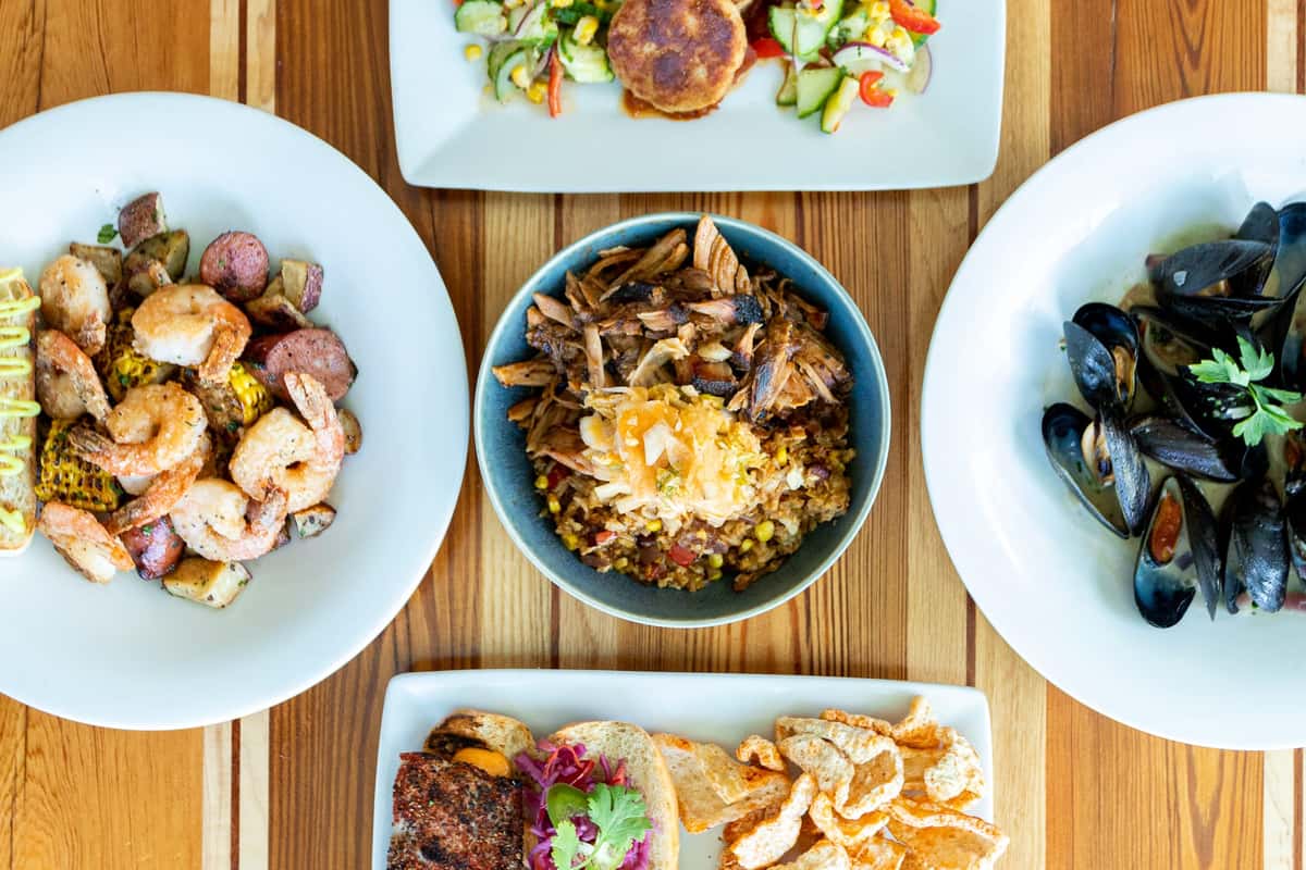 The image presents an overhead view of several beautifully plated dishes arranged on a wooden table, including seafood, salads, and hearty entrées. The vibrant presentation highlights the variety and fresh flavors offered at Backstreet Grille & Tavern.