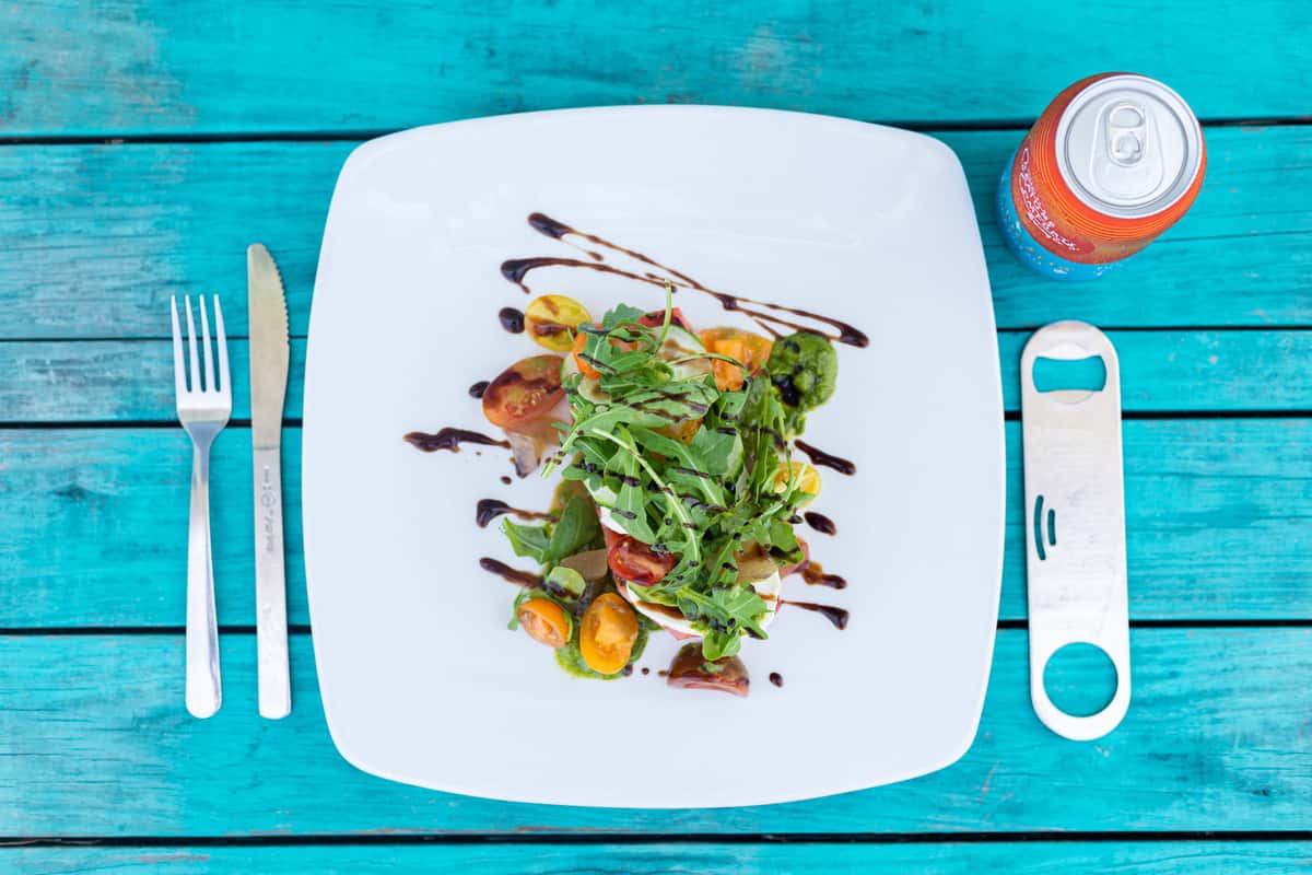 The image features an elegantly plated seafood entrée garnished with fresh greens and herbs, presented on a white square plate atop a vibrant turquoise table. The clean presentation and colorful accents create a fresh, coastal-inspired dining aesthetic.