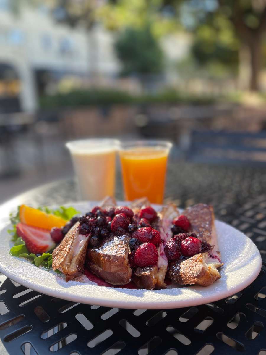 A beautifully plated breakfast features thick slices of French toast topped with a generous mix of fresh berries, including blueberries and raspberries, finished with a light dusting of powdered sugar. On the side, there's a colorful assortment of fresh fruit. The dish is accompanied by a glass of orange juice and a cup of coffee, creating a warm and inviting morning meal setting.