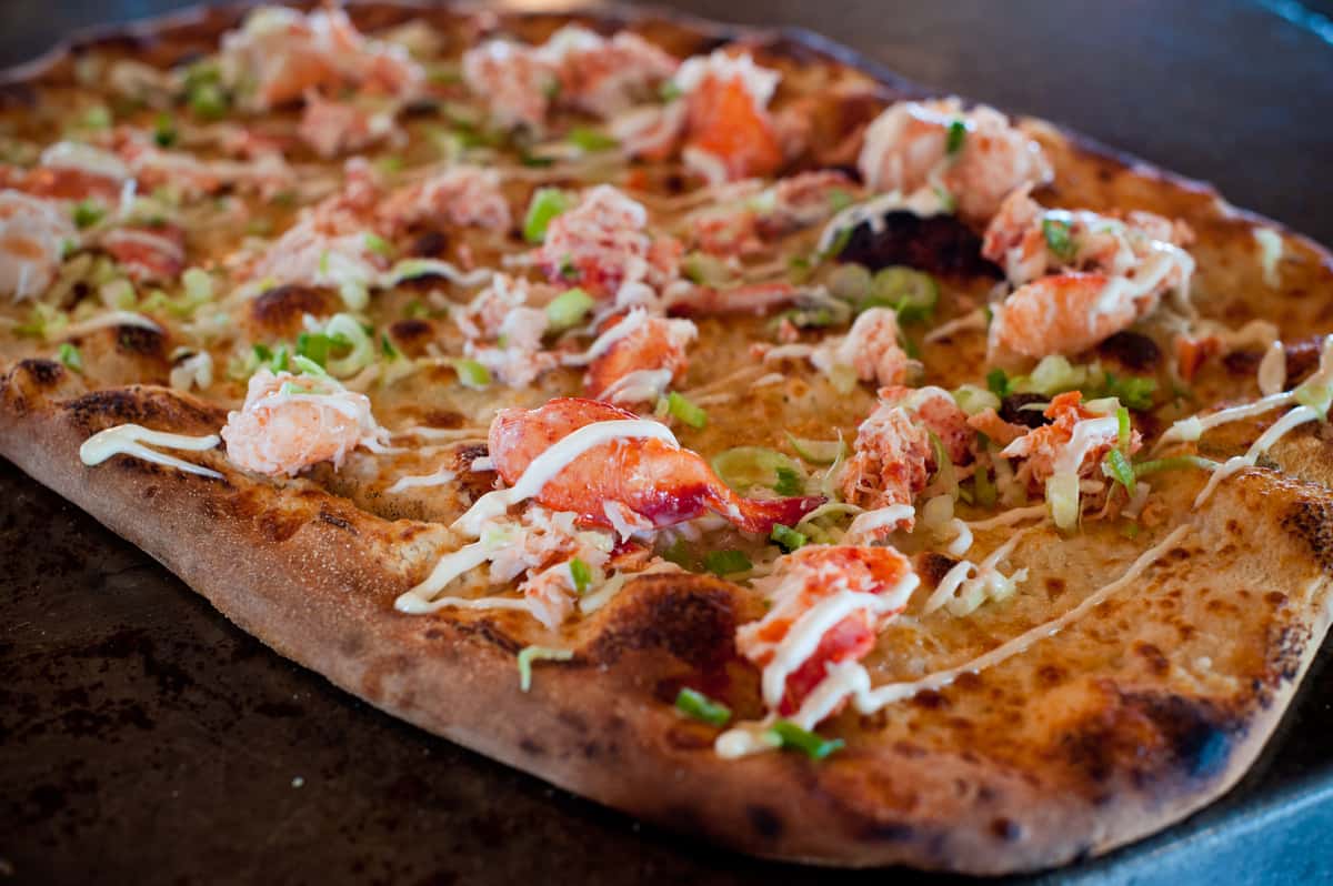 Wood-fired flatbread pizza topped with chunks of lobster, green onions, and creamy drizzle on a crispy charred crust