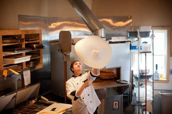Chef tossing pizza dough in a commercial kitchen with wood-fired oven and stainless steel prep area