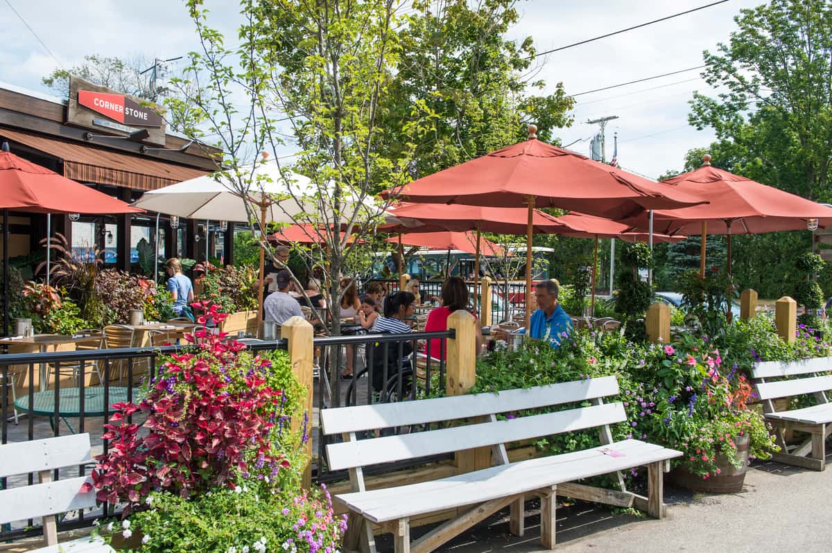 Outdoor restaurant patio with red umbrellas, garden seating, and guests dining in a lush, casual setting