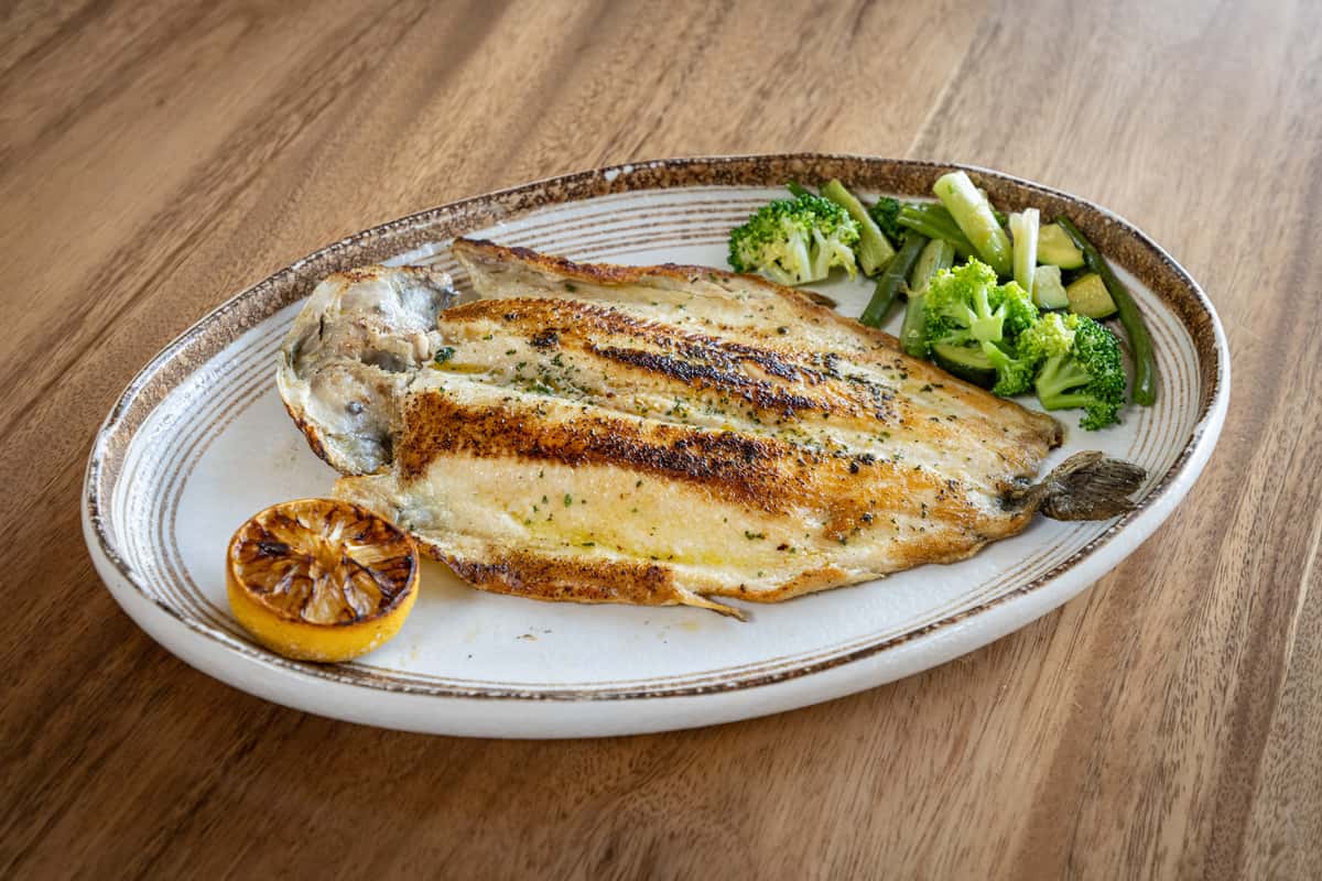 A whole grilled fish (likely branzino or sea bass) served on a white plate with a side of broccoli and grilled lemon.