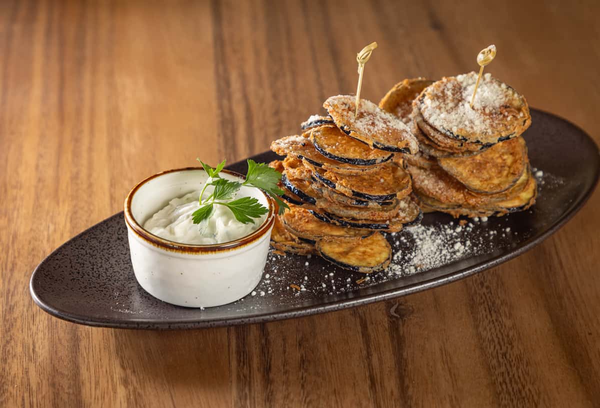 A stack of crispy, breaded fried zucchini or eggplant rounds served with a white dipping sauce