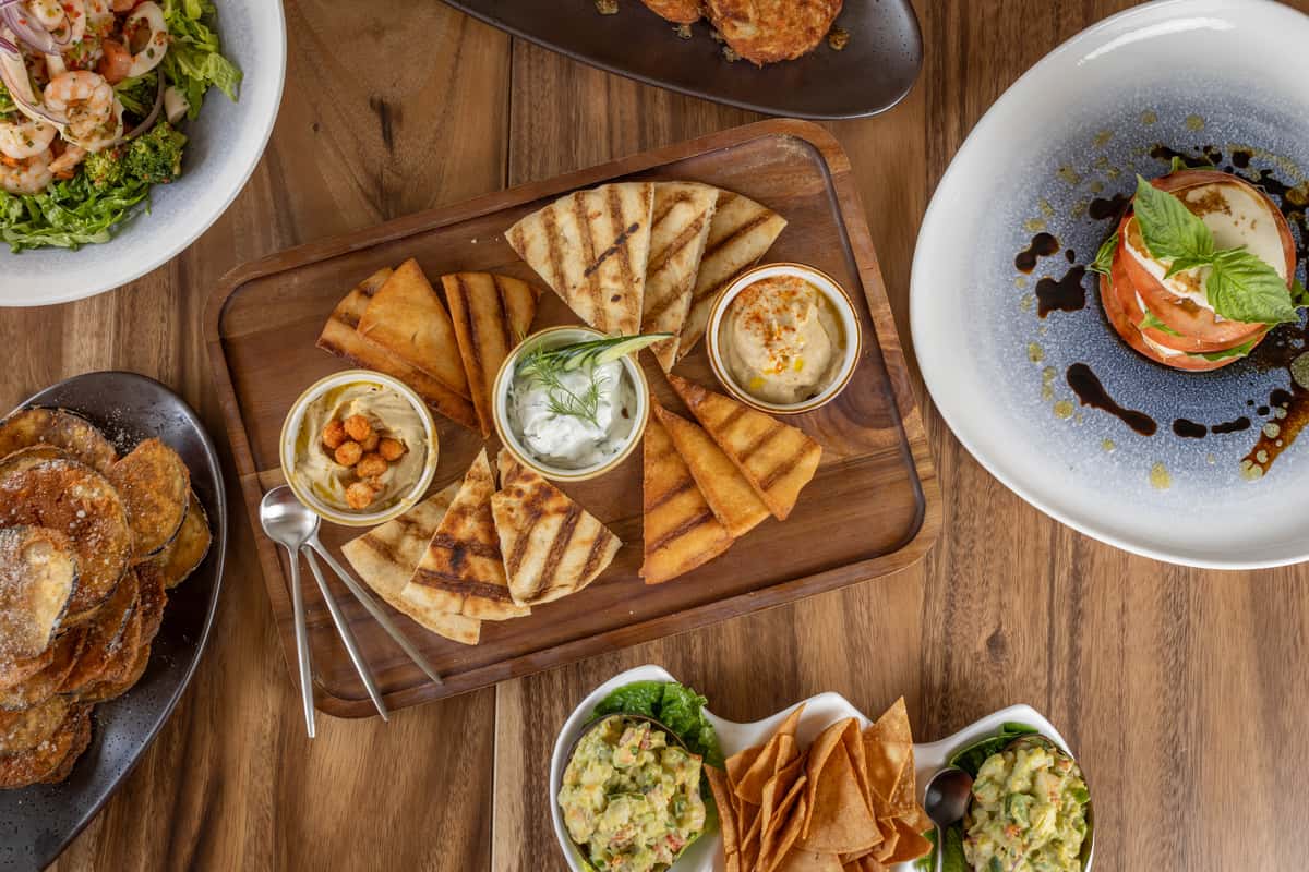 An overhead view of a wooden board with pita bread triangles and bowls of hummus and tzatziki.