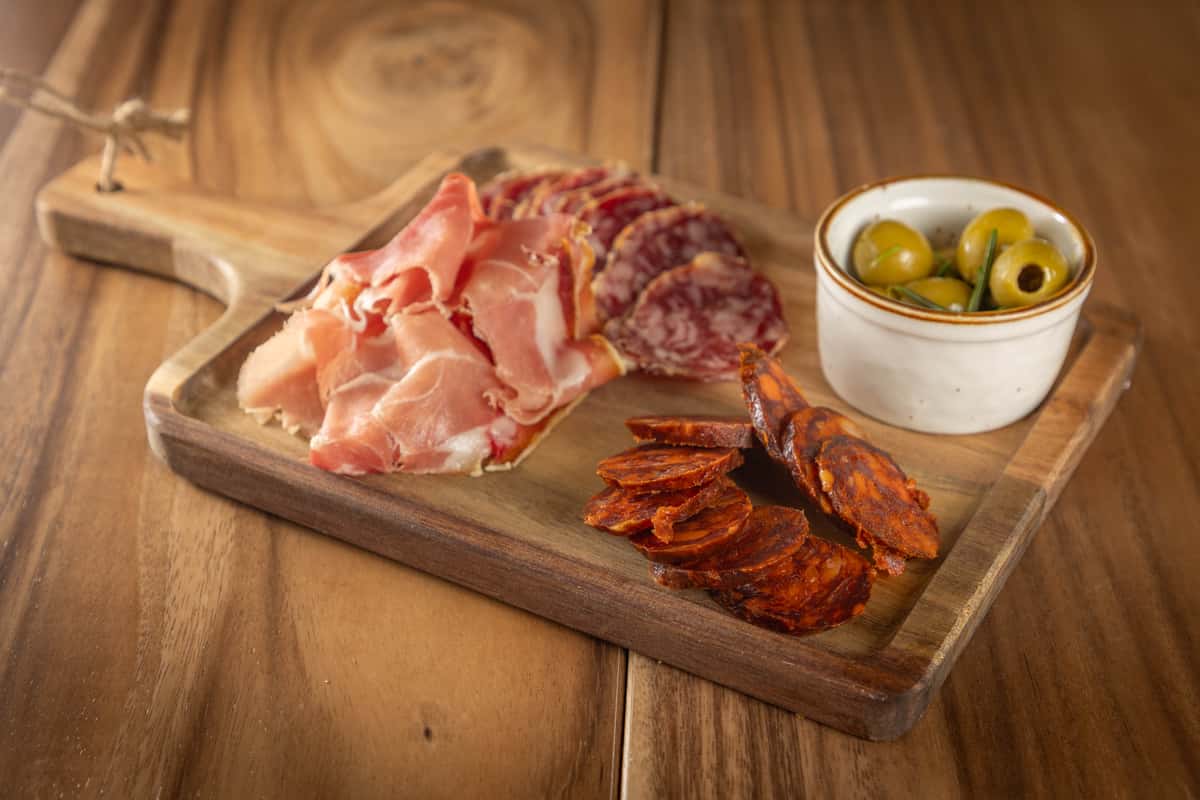 A wooden board displaying cured meats (prosciutto, chorizo) and a small bowl of olives.