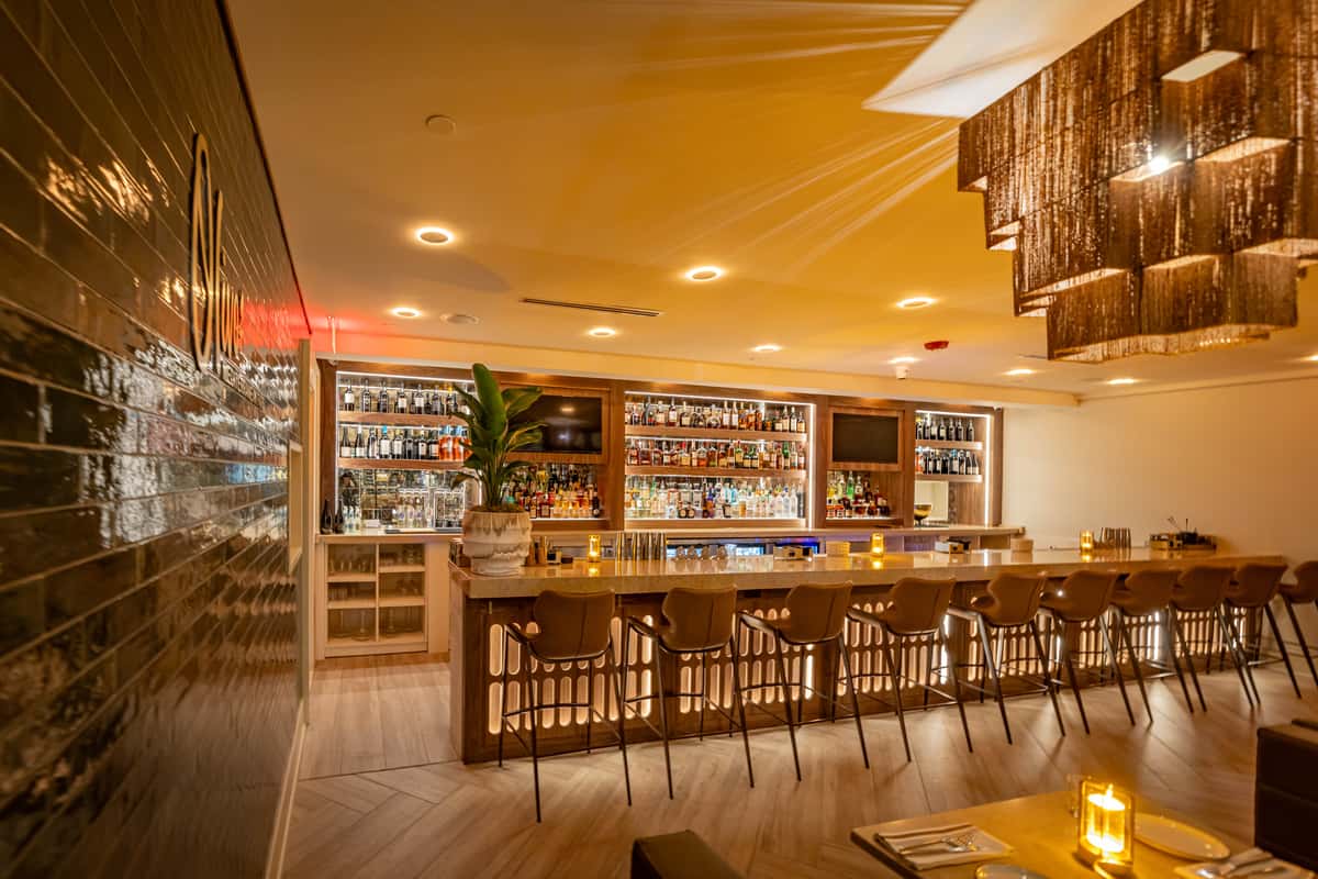 A wide interior shot of a restaurant bar area featuring wooden stools, shelving, and warm lighting.