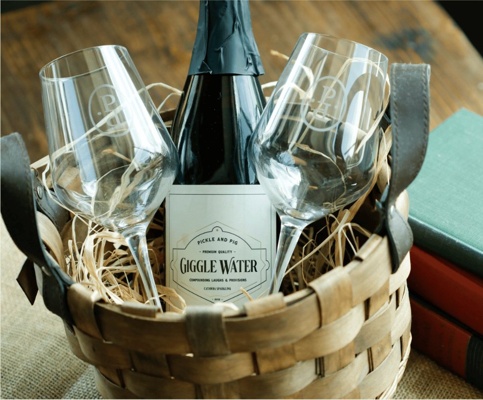 Rustic gift basket featuring a bottle of "Giggle Water" sparkling wine from Pickle and Pig, paired with two engraved wine glasses on a bed of straw, set on a wooden table with vintage books in the background.