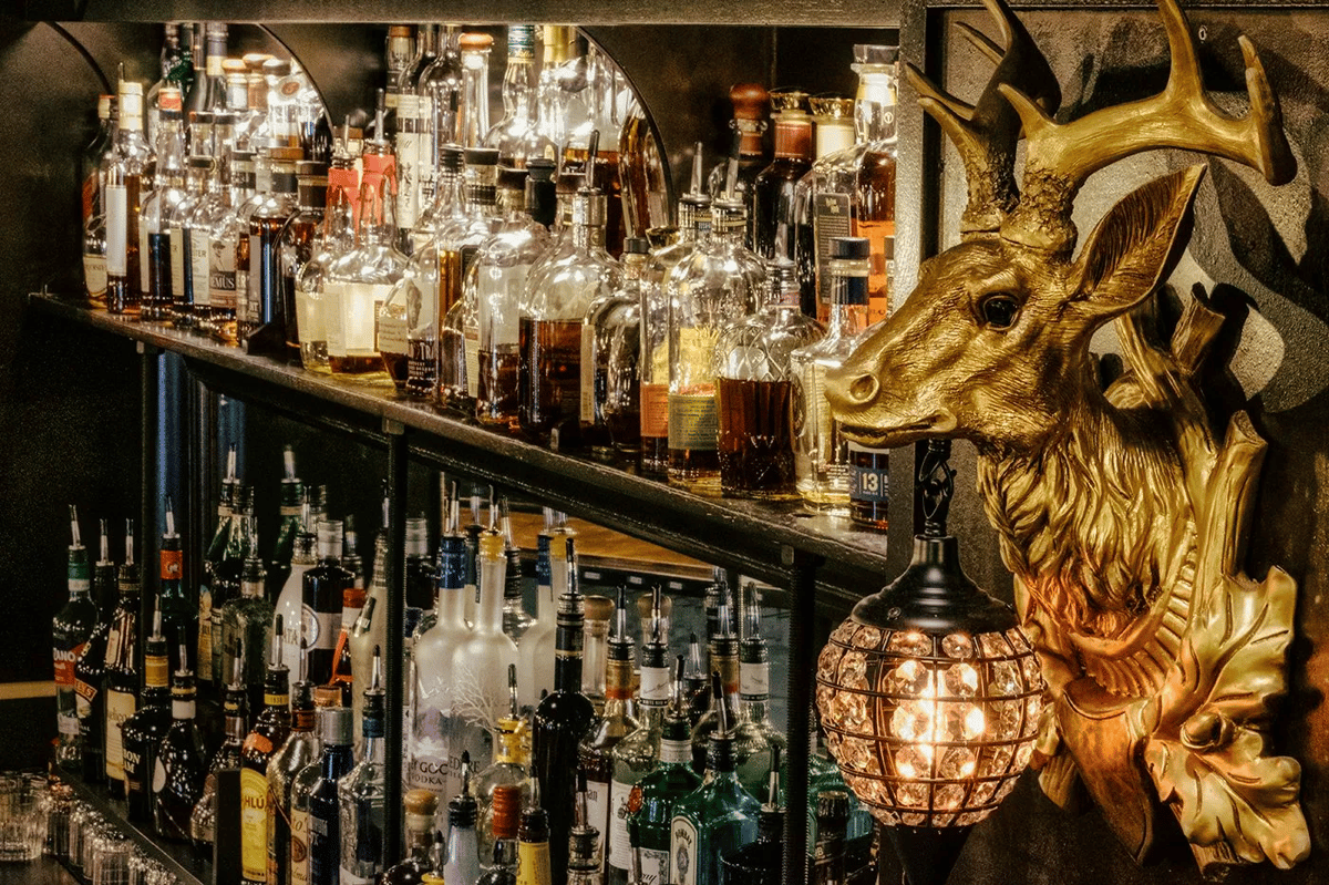 Upscale bar display with a wide selection of liquor bottles arranged on mirrored shelves, accented by a golden deer head wall mount and a warm glowing pendant light for a rustic yet elegant ambiance.