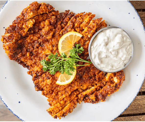 Crispy fried cutlet served with lemon slices, parsley garnish, and a side of creamy dipping sauce on a rustic white plate.