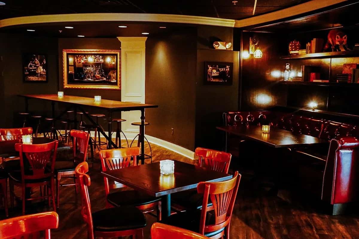 Cozy restaurant dining room with dark wood tables, red chairs, and leather booth seating, softly lit by candles and wall sconces, creating a warm and intimate atmosphere.